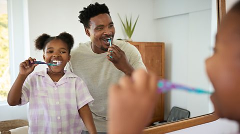 A father and daughter brush their teeth together whilst standing by a mirror
