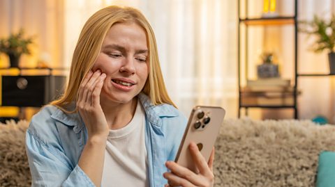 A woman holds her jaw in discomfort whilst looking at her phone