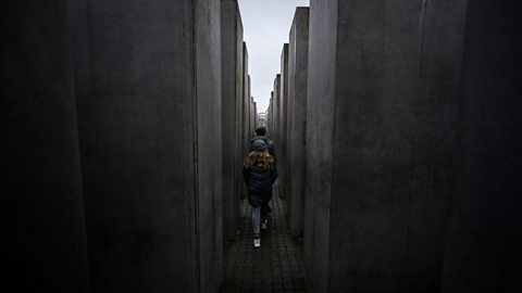 Visitors walk through narrow concrete corridors formed by tall stone slabs at the Holocaust Memorial in Berlin.