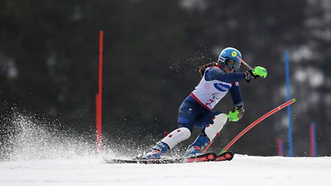British visually impaired skier Menna Fitzpatrick on her way to gold in a slalom run. Her guide is out of shot, she is taking a left turn past a red pole. She is wearing a blue skin suit and helmet, with white bib and shin guards