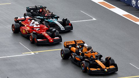 Three Lego versions of Formula One cars are shown on a track. The front car is an orange and black McLaren, driven by Lando Norris and Oscar Piastri. It's followed by a red Ferrari driven by Charles Leclerc and Lewis Hamilton, then a teal and black Mercedes, driven by George Russell and Kimi Antonelli