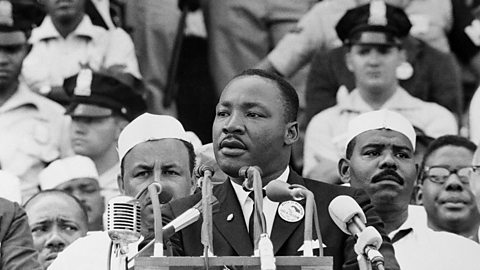 A historic black‑and‑white scene shows Dr Martin Luther King, Jr delivering a major civil rights address from a podium surrounded by microphones, with a large crowd gathered behind.