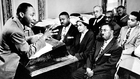 A black‑and‑white photograph shows Dr Martin Luther King Jr. addressing a small seated audience inside a meeting room, this audience includes Rosa Parks and people in suits listening attentively from folding chairs arranged in rows.