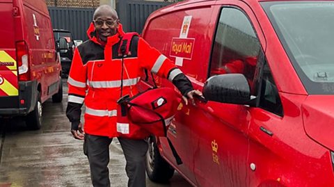 A postman David Bostock stands next to his Royal Mail van wearing a matching red uniform winter coat, trousers and big boots