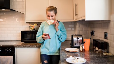 Girl eats toast while scrolling on smart phone in kitchen