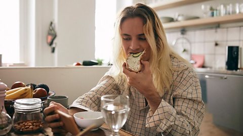Young person eating cream cheese and cucumber on bread, a bowl of cereal while watching their phone