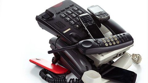 A pile of old phones sit on a white table. At the bottom of the pile is a large, black landline machine with buttons and no phone resting on it and scattered on top of various mobile phones and smartphones with touch screens