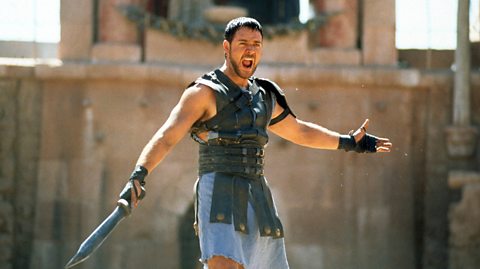 Russell Crowe in Gladiator. He has short brown hair and a beard and is screaming in the photo with his arms opened out. He wears leather armour and holds a sword in his right hand