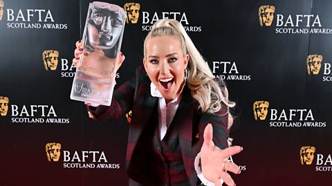 Sheli McCoy poses with the Audience Award for Gladiators during the 2025 BAFTA Scotland Awards. She has a tartan red suit on and her long, blonde hair in a ponytail. She smiles at the camera with the transparent glass BAFTA Award, which looks like a head, in her right hand. Behind her is a black advertising board on the red carpet promoting the BAFTAs