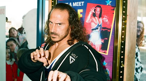 Wolf, Wolf, real name Michael van Wijk, at a Gladiators event in Blockbuster. He has long, dark curly hair, a moustache and beard and has an intense look on his face as he poses for the camera. His hands are posed like claws and he wears a black hoody jacket which says Wolf on it. Behind him is a poster of himself in the same pose, wearing his metallic blue Gladiator uniform. At the window behind him, fans peer through to catch a look at the Gladiator
