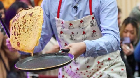 A person wearing a patterned apron and bowler hat flips a large pancake from a frying pan during a lively indoor event, with onlookers gathered in the background.