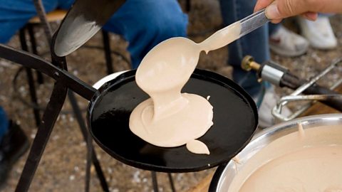 A person pours pancake batter onto a small round griddle using a ladle, with cooking equipment and a bowl of batter visible in the outdoor setting.