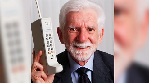 Dr. Martin Cooper with the first portable handset. He holds the large, brick shaped grey phone with an antenna in his right hand while smiling. He has white hear and a white beard and wears a navy blue suit jacket, dark blue tie and light blue shirt