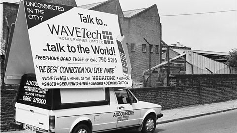 A black and white photograph of a car carrying advertising boards for 1980s mobile phones, WaveTeck and Vodafone. It is parked on the side of a street with houses behind and the board rests on top of the car