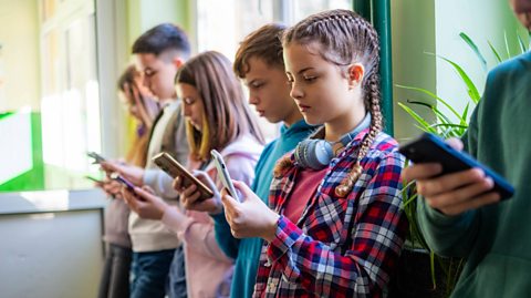 In a corridor with plants resting on windowsills and the sunlight beaming in, boys and girls around the age of 12 stand lined up, all on their phones. They are in their own clothes and are all focuses on what they are watching on their touchscreen smartphones