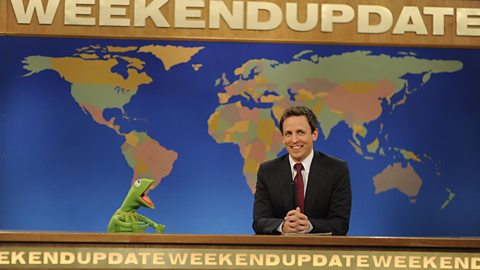 Kermit the Frog appears behind the desk of Weekend Update on Saturday Night Live alongside anchor Seth Meyers. The backdrop is a map of the world with the words Weekend Update appearing prominently