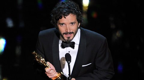 Bret McKenzie, wearing a black tuxedo and bow tie, leans towards the microphone while clutching an Oscar award