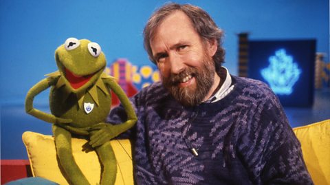 Kermit the Frog wears a Blue Peter badge sat next to Jim Henson on a sofa with yellow cushions. A neon Blue Peter sign is visible in the background