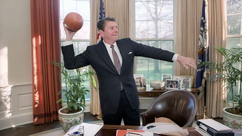 Ronald Reagan holds an American football in his right hand, posing as if to throw it, while stood behind his desk in the Oval Office at the White House