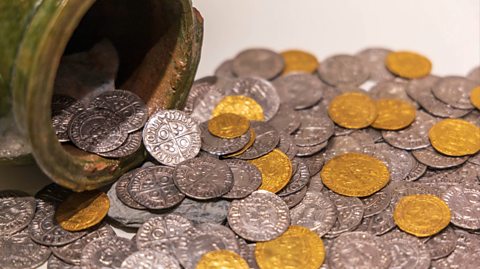 An old clay pot lies on a white table as a pile of coins spills out. The coins are ancient, the majority silver and others gold and thin.