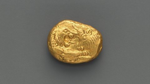 A Lydian Lion coin lies on a grey table. It is thick, gold and round, with a lion and bull illustration embedded into it