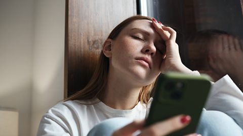 teen girl holds head as if head hurts from scrolling on her mobile phone