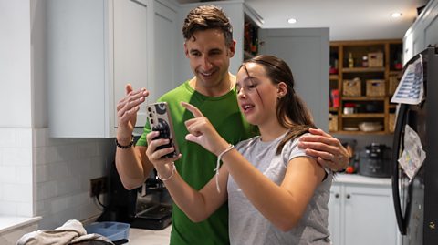 father and daughter share a joke over a mobile phone screen in the kitchen 