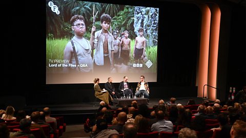 Emma Bullimore, Marc Munden, Jack Thorne and Joel Wilson speak during a Q&A at a special screening of BBC's Lord Of The Flies. They sit on a dark stage with an audience seated in red chairs. Behind them on the big screen is a photos of children on set from the series