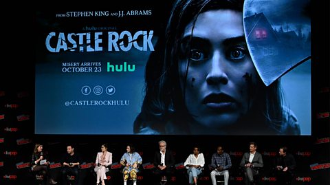 Dustin Thomason, Lizzy Caplan, Tim Robbins, Paul Sparks, Yusra Warsama, Barkhad Abdi, Matthew Alan and Elsie Fisher speak on stage during J.J. Abrams and Stephen King’s Castle Rock season two world premiere. They sit on stage in a row seated on red chairs. Behind them is a large screen with a promotional photos of one of the lead characters for the series