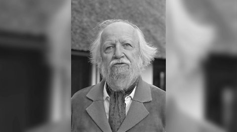 A black and white photo of author William Golding. He has a white beard and white flicky hair and wears and suit jacket and neck scarf as he stands outside his house