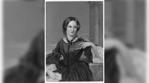 A black and white portrait of Charlotte Brontë. She smiles as her hair is in a centre part and a low bun. She wears a dark, long-sleeved dress with white ruffles on the neck and sleeves and has her left arm rested on her lap, while her left elbow rests on a side table
