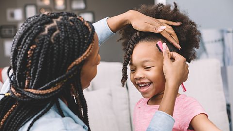 A mother with braided hair and her back to the camera, styles her daughter's hair. Her daughter has afro hair.
