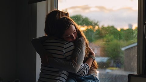 Mother comforts her daughter in an upstairs bedroom silhouetted against a sunset through the window