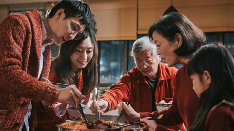 A family, a little girl, her mum and dad and grandmother and grandfather, all wear read and surround a dinner table, digging into food in front of them