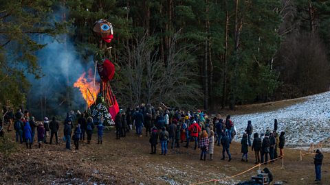 A large red and brown effigy is set alite as part of the Užgavėnės celebrations. It takes place near a forest, with some snow on the ground.