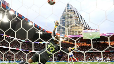 View from behind a goal net as Chloe Kelly aims a penalty into the roof of the net beyond the outstretched arm of Spanish goalkeeper Cata Coll in the Euro 2025 final
