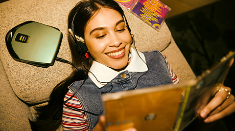 A young woman with long dark hair lies on a cream bed wearing a red and white striped long-sleeved top under a denim waistcoat. She smiles as she reads inside of a CD cover and has headphones in connected to a small portable CD player