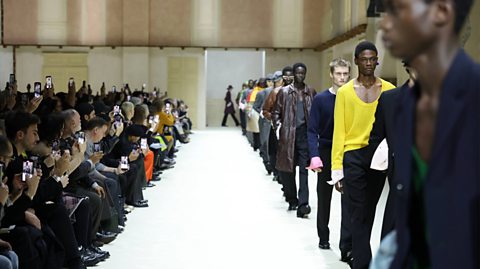 Model walks the runway at a Prada fashion show during the Milan Fashion Week. Male models wear various clothes as they walk down a white runway. To the left sits an audience, with many members taking photos on their camera phones