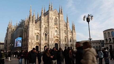 The Duomo di Milano - a large, white Gothic cathedral, sits in the middle of a busy square. Around it are other buildings and crowds of tourists taking photos and looking at the view