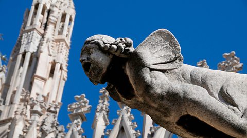 A stone gargoyle sticks out at Milan Duomo, a large Gothic white cathedral. The gargoyle has an open mouth, curled hair and wings