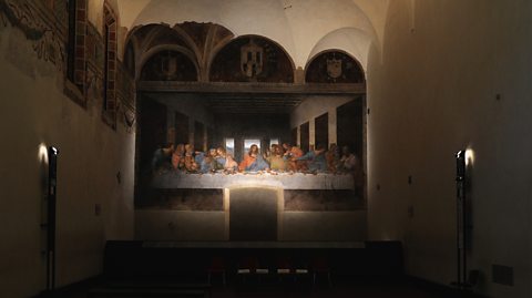 The Last Supper on display in a dark room. The image depicts Jesus at the centre of a dinner table with his 12 disciples either side and their reactions as he tells them he will be betrayed