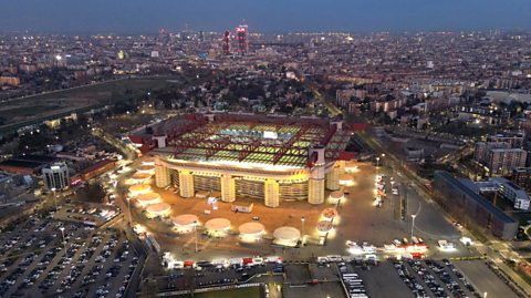 An aerial view of The Stadio Giuseppe Meazza at night. Around the stadium you see buildings across the city. The stadium is lit up and through the roof, you can see the green pitch. The stadium is in the shape of a square with curved edges