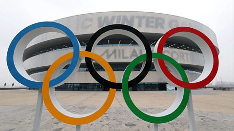 The Olympic rings outside the Milano Santagiulia Ice Hockey Arena. The arena behind is white and grey and the sky is white and foggy. There are five Olympic rings intertwined - one blue, one yellow, one black, one green and one red