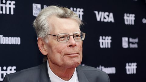 Stephen King smiles for the camera wearing a grey suit and white shirt. He has dark square shaped glasses and grey hair