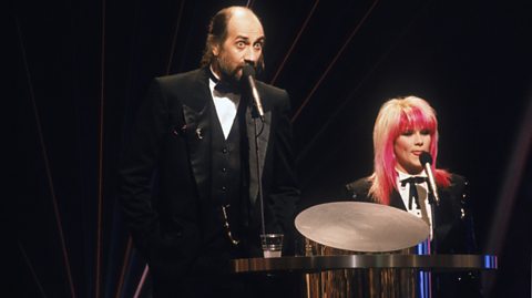 Mick Fleetwood and Sam Fox stood in front of microphones at the 1989 Brit Awards. Both are wearing tuxedos.
