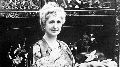 A black and white photo of author Agatha Christie. She sits at a table surrounded by flowers and wears a floral silk robe with her curly hair tied up in a bun