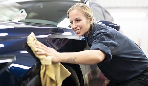 An image of a girl washing a car, she is wearing overalls and using a yellow cloth. The car is dark blue.