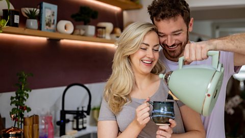 A man pours a cup of tea from a kettle for his partner in the kitchen.