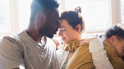 A young couple embrace, a toddler in the mother's arms.
