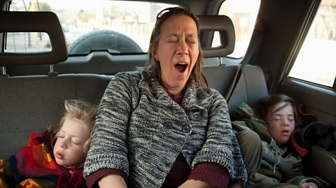 mother yawns with exhaustion as her young children sleep beside her on the back seat of a car